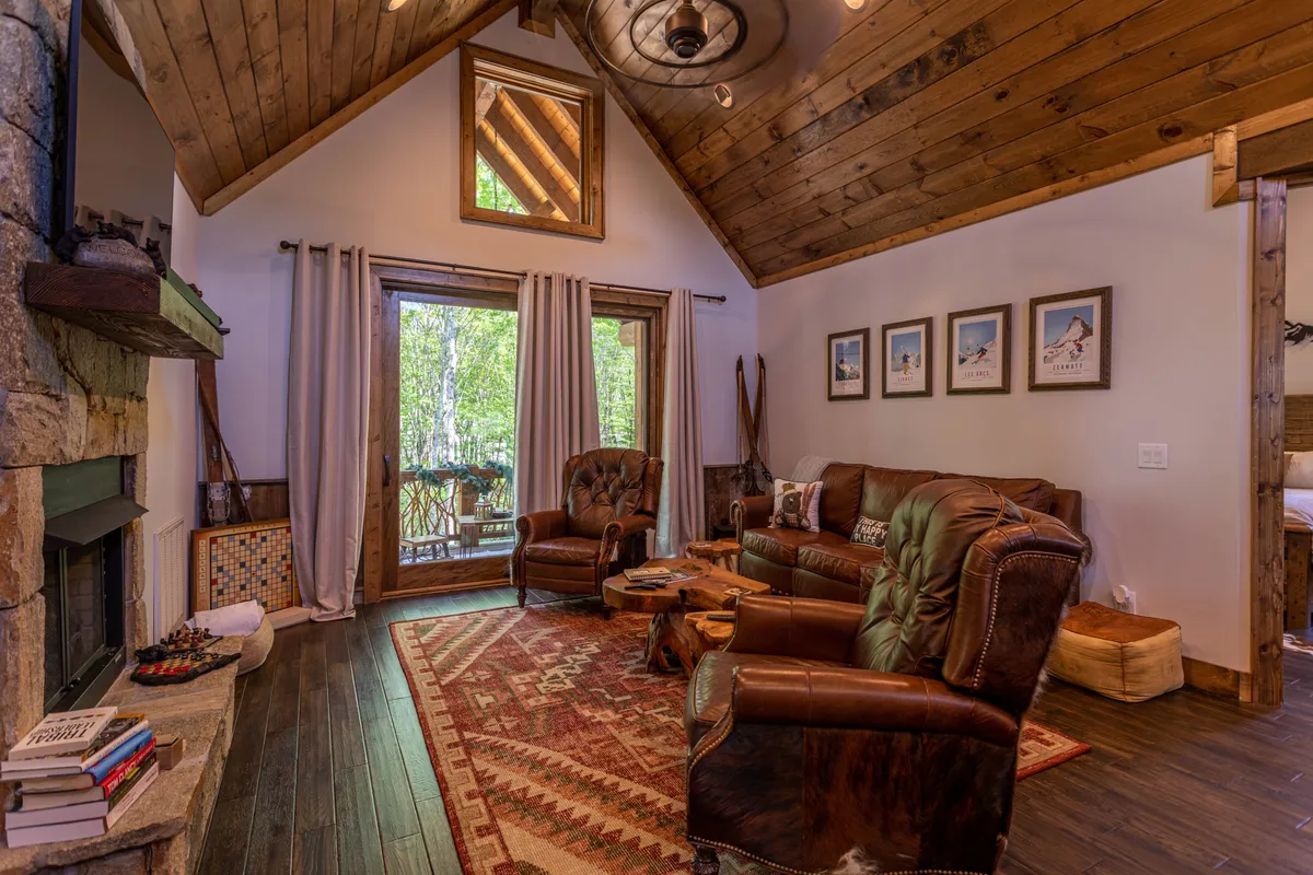 A comfortable lodge living area featuring a large stone fireplace, exposed wooden beams, cozy seating, and a central rug. The room offers an inviting atmosphere for family gatherings and relaxing together after a day of resort activities.