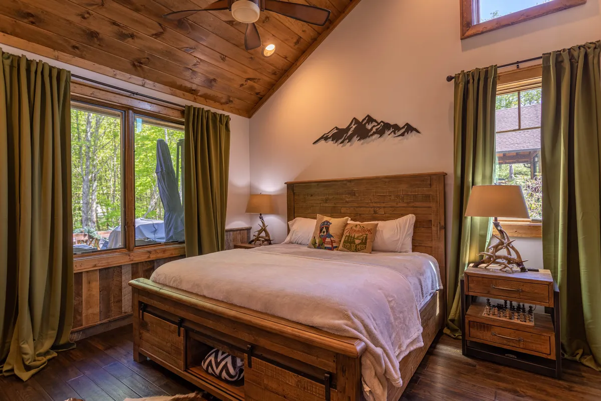 A serene bedroom featuring a wooden bed frame topped with soft, decorative pillows. Mountain-inspired wall art hangs above the headboard, and large windows framed by green curtains invite natural light and tranquil views of the outdoors, creating a peaceful, airy atmosphere.