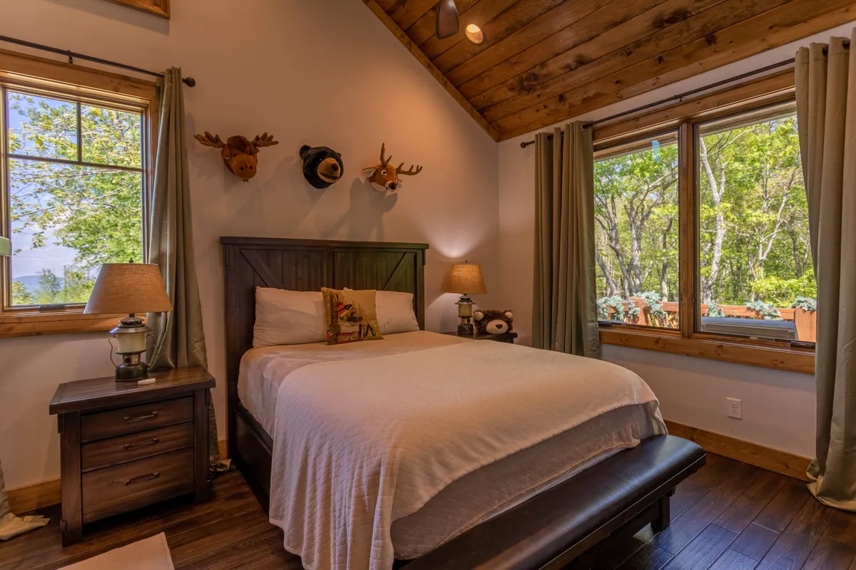 A warmly lit bedroom with a cozy bed positioned near a large window, letting in natural light and offering a glimpse of the outdoors. The room features inviting wood accents, mounted animal heads, and a nightstand with lamps, creating a comfortable retreat atmosphere ideal for relaxation after a day of winery adventures.