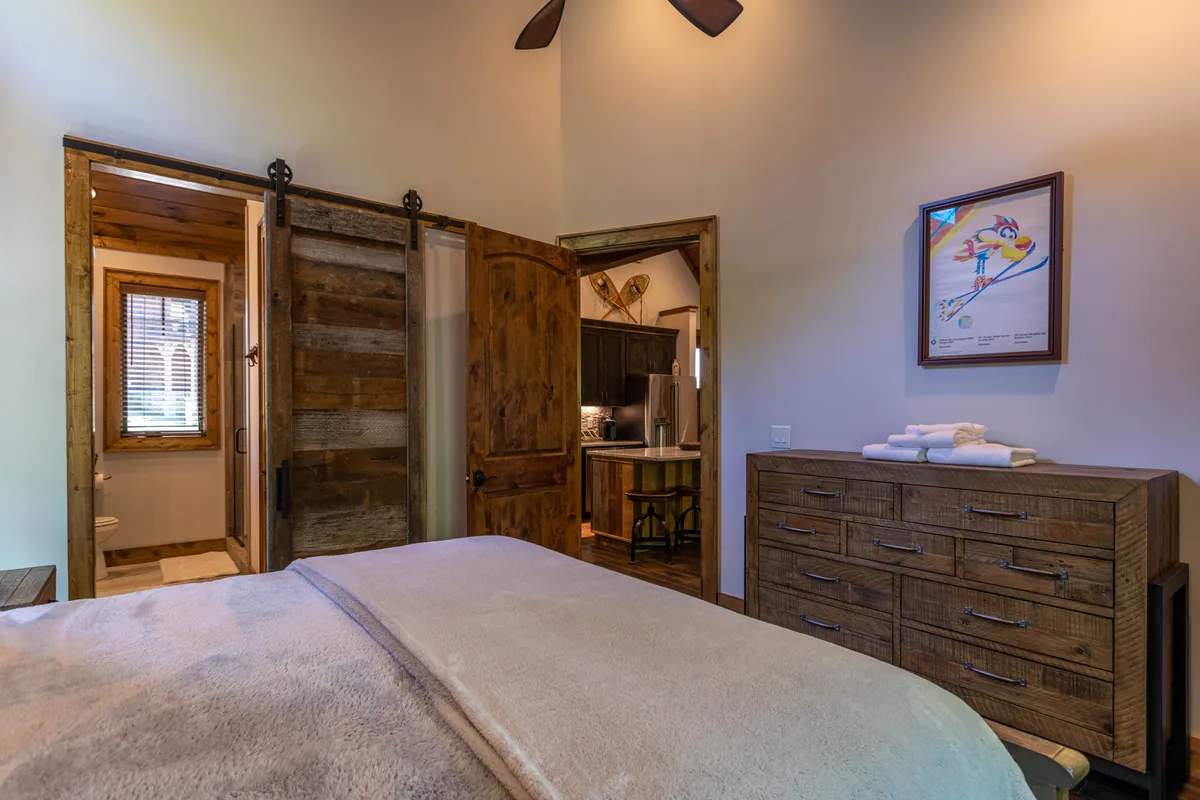 A cozy bedroom with plush bedding, rustic wooden sliding doors, a sturdy dresser, and tasteful wall art. Gentle natural light illumes the soft textures, evoking comfort and a welcoming, homey vibe.