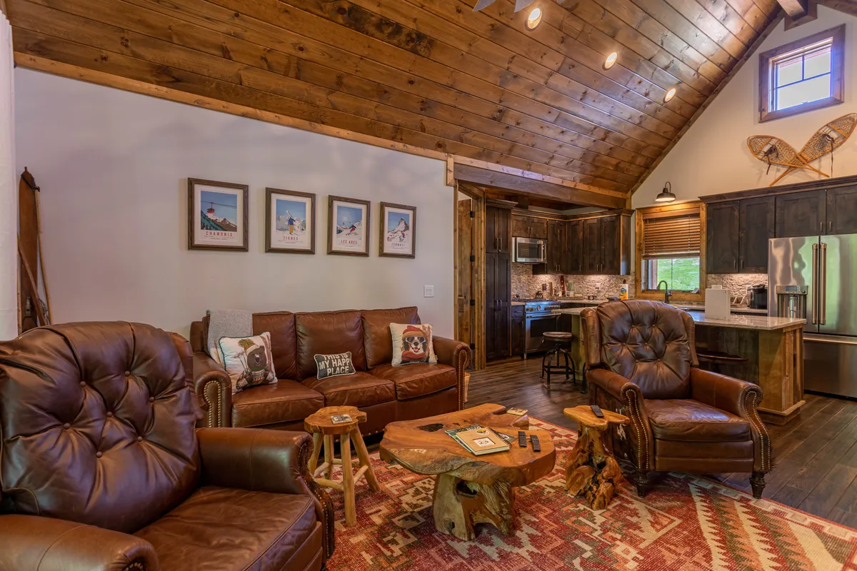A cozy living room scene with leather chairs, a wooden coffee table, and clear sightlines into the adjacent kitchen area. Framed artwork and warm lighting add character, creating a comfortable space for relaxing, playing games, or entertaining.