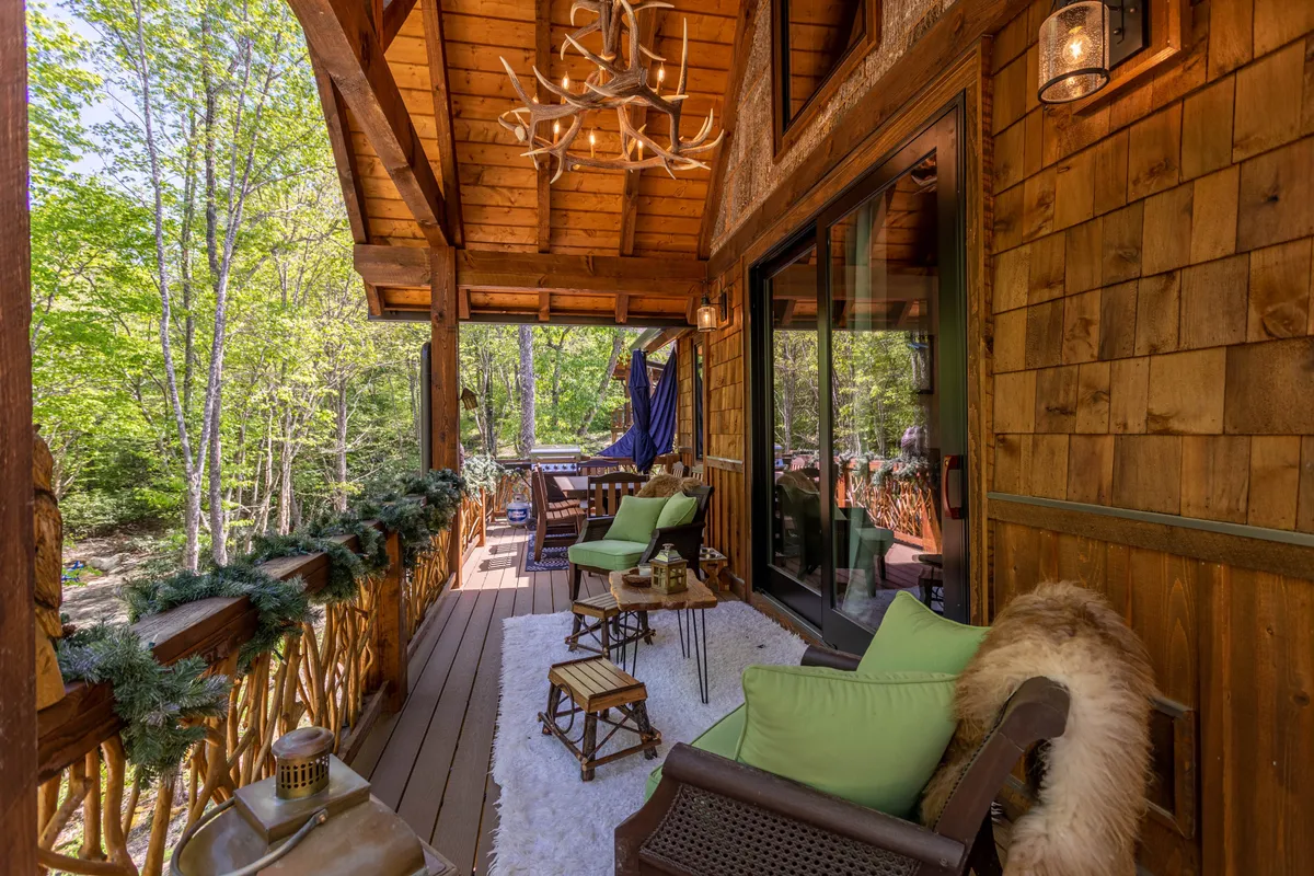 The image showcases a cozy wooden deck adorned with plush green cushions, a rustic antler chandelier, and a soft rug—framed by sturdy wooden railings and surrounded by serene mountain nature. The setting invites relaxation, morning coffee, and evening gatherings in a welcoming, nature-inspired atmosphere.