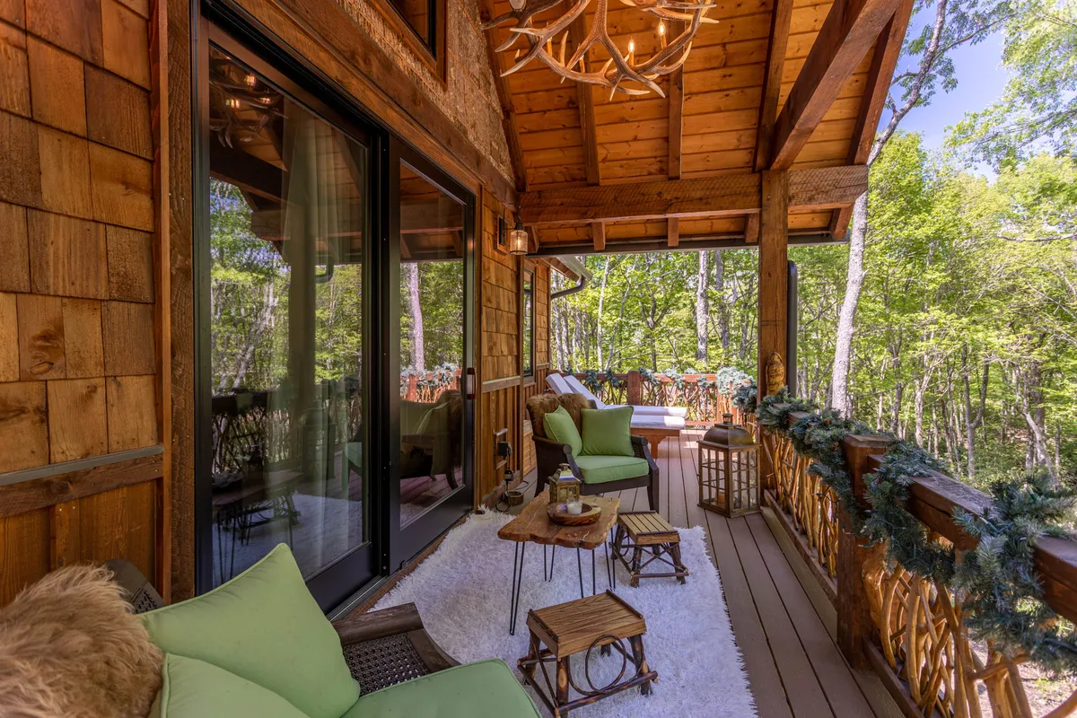 A comfortable outdoor seating area on a wooden deck, framed by natural wood finishes and surrounded by lush green trees. The tranquil setting provides a cozy spot for relaxation, with ample seating and soft lighting that hints at gatherings that extend from sunrise to evening.