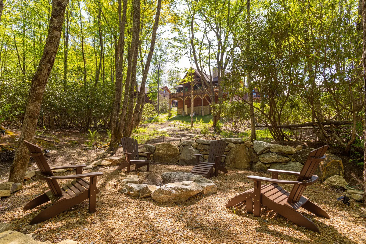 Wooden chairs encircle a glowing fire pit beneath a canopy of tall trees, with a distant glimpse of the house. The scene evokes communal warmth, storytelling, and laughter, all set against the backdrop of a private, wooded lot.
