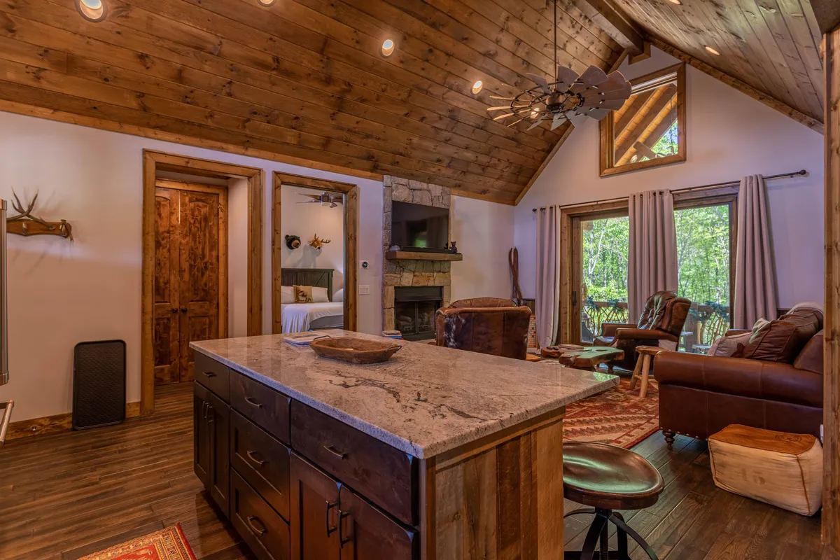 A spacious, warm open-concept area featuring a modern kitchen island, a cozy stone fireplace, large windows with views of the outdoors, and a comfortable seating area. The room is filled with natural light and creates an inviting hub for family gatherings.