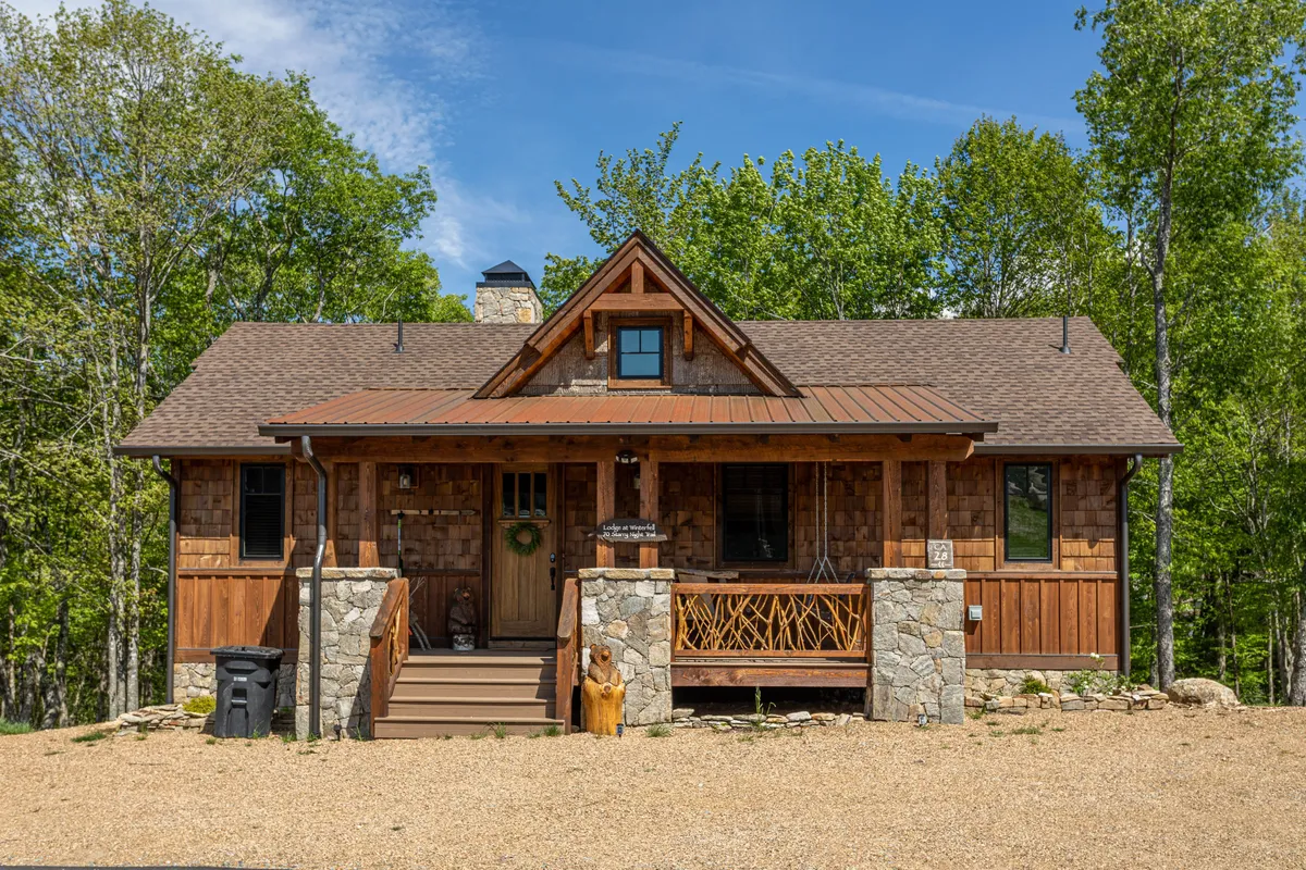 The photo shows the lodge’s wooden front door and stone foundation set against a backdrop of tall trees, highlighting the seamless transition from indoor comfort to outdoor adventure. The image evokes a sense of welcome and hints at the surrounding woods and opportunities for exploration right from the doorstep.